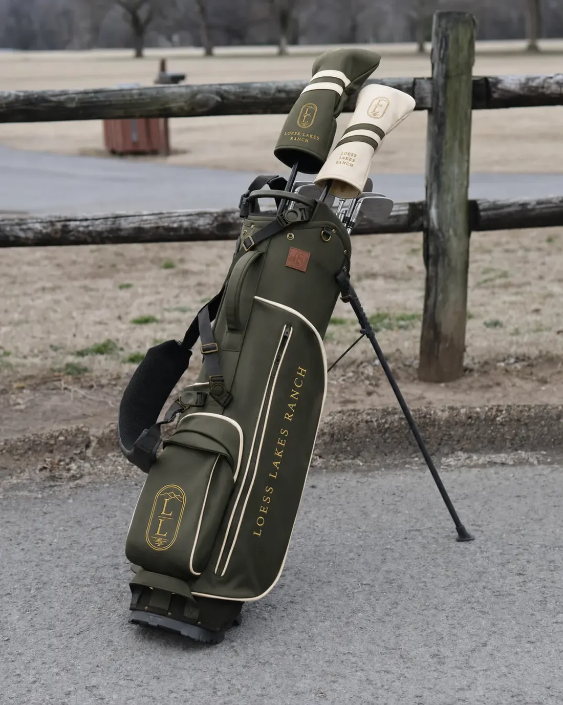 Olive LOESS LAKES RANCH stand bag with cream piping, antique-gold lettering and two branded headcovers, resting against a split-rail fence