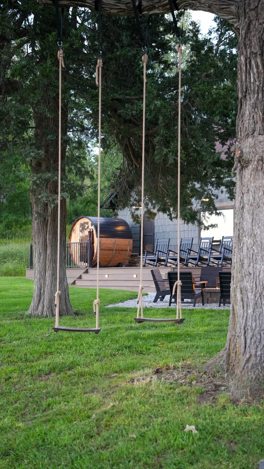 Two rope swings beneath mature trees with the cedar sauna, deck and firepit beyond