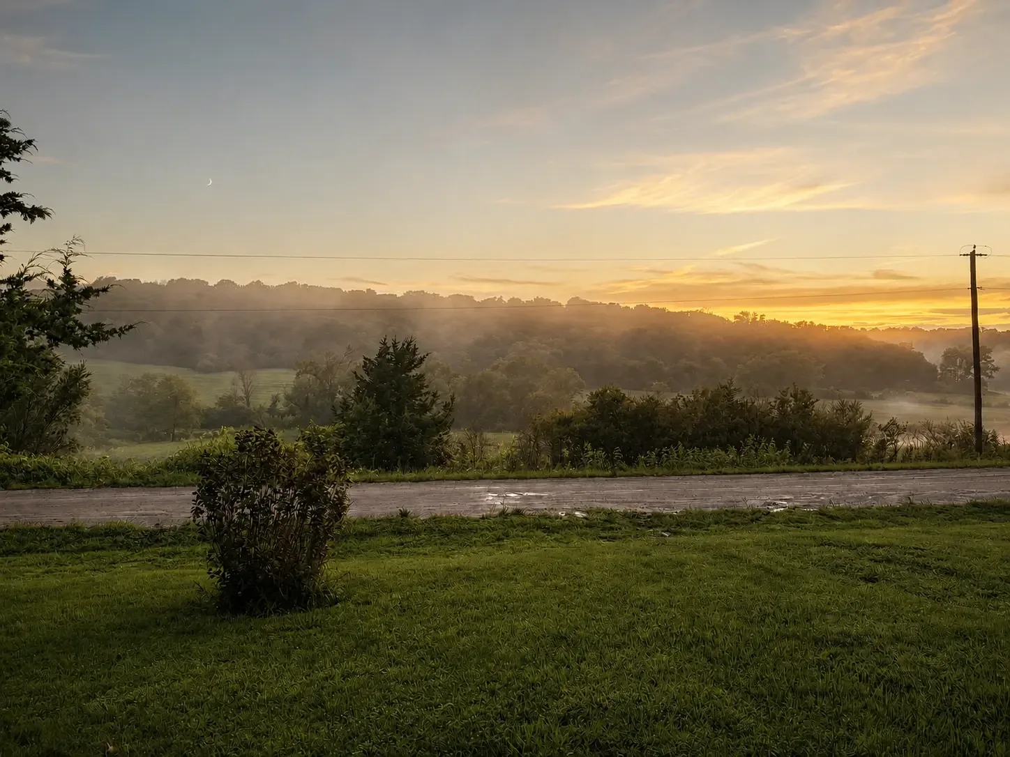 Mist rising over rolling hills at sunrise with a crescent moon above the ranch road
